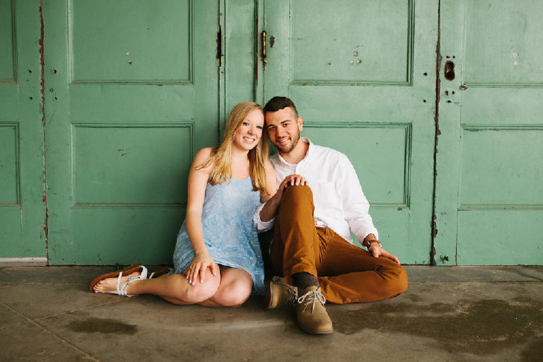 Couple smiles together as they look at camera while sitting in front of large green doors at Asbury Park