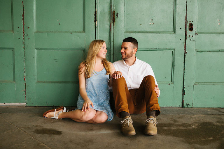 Fiances look in love at one another sitting in front of rustic green doors for engagement photography