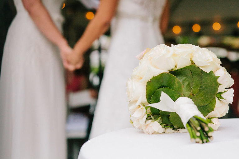 lesbian couple holding hands in the background with focus on the bridal bouquet of white roses
