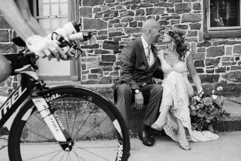 bride and groom sit with bicycle for summer wedding portraits outside a historic stone building near the Lumberville foot bridge over the Delaware River by Kasey Ivan Photography outside the Black Bass Hotel near New Hope PA