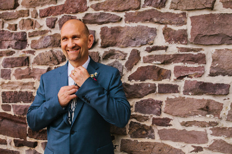 English groom in a blue suit adjusts his tie outside a historic stone building on the Delaware River by Kasey Ivan Photography