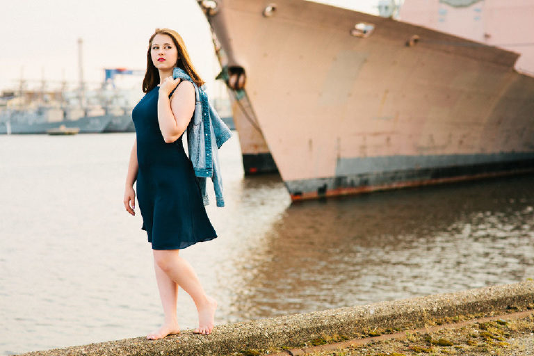 Senior posing walking on the edge of the water with jean jacket over the shoulder at the navy shipyard in philadelphia posing for Kasey Ivan Photography