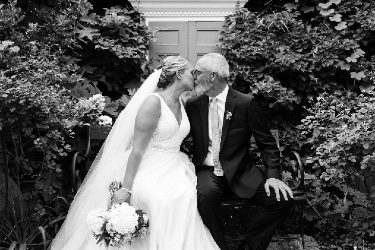 Black and white picture of a bride and groom kissing on a bench in a rustic garden in the Porches on the Towpath in New Hope and Lambertville
