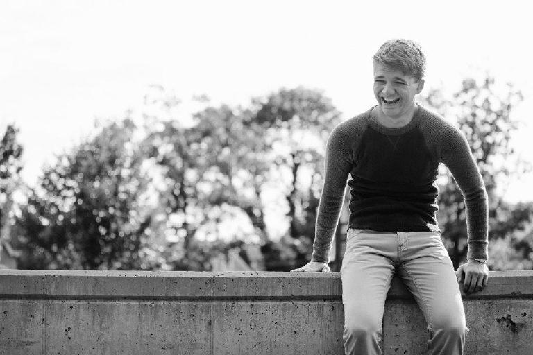 high school boy sits on ledge and laughs for senior pictures